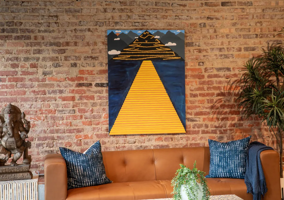 A geometric pyramid painting by Micah The Artyst hung above a leather sofa against a brick wall