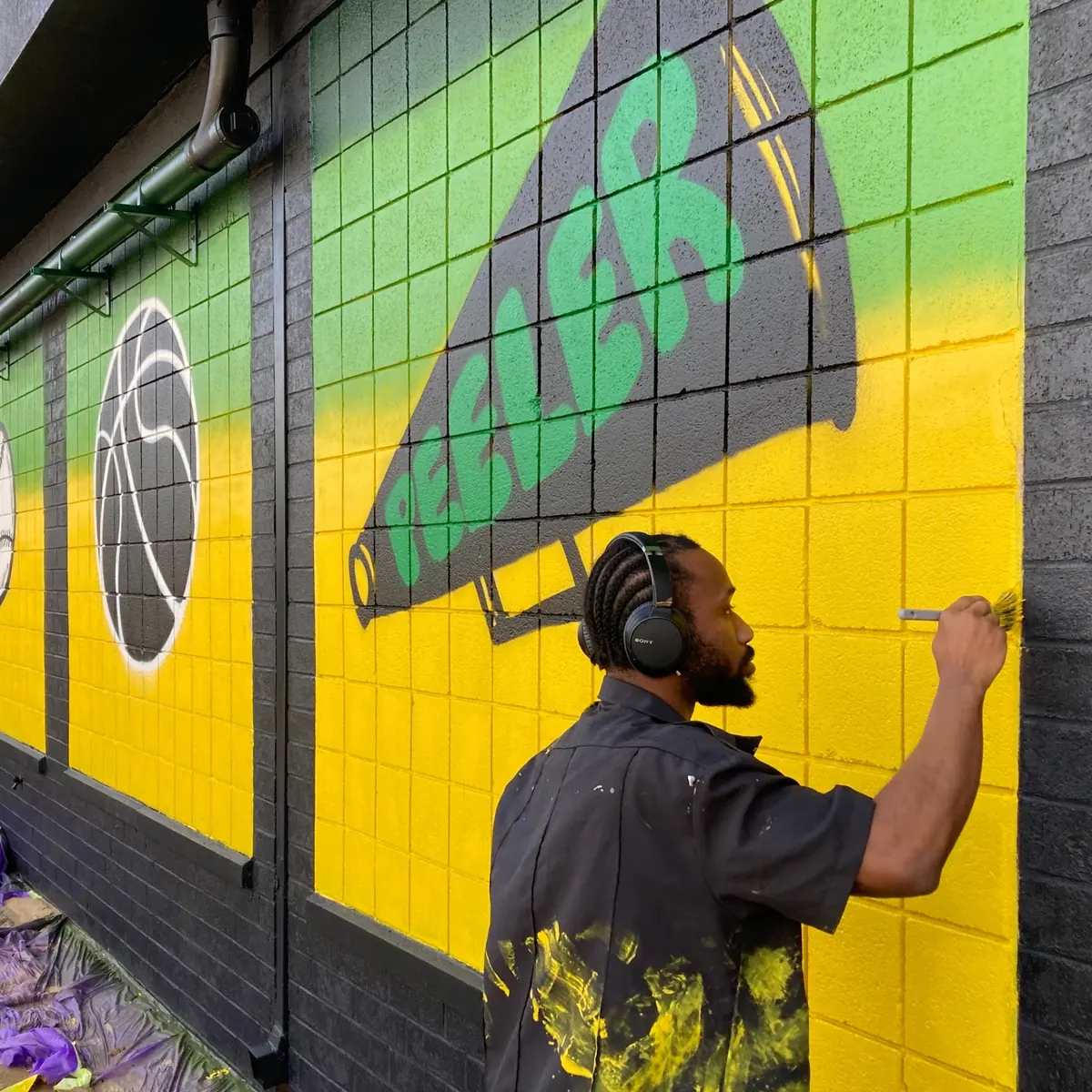 Micah The Artyst painting a green and yellow megaphone mural on a block wall